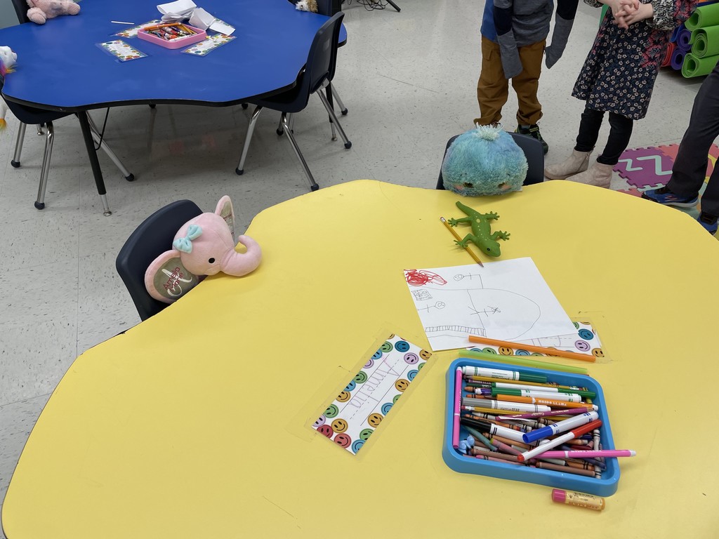  A high-angle shot of a yellow, clover-shaped classroom table. A pink stuffed elephant with a bow sits in a chair on the left, and a round, teal fuzzy stuffed animal sits in a chair at the top. On the table, there is a green plastic lizard, a drawing on white paper, a blue tray filled with markers and crayons, and a name tag that reads "Amelia." In the background, a blue table and the legs of children are visible.  Photo 2 (IMG_6094.jpg) A yellow, clover-shaped table in a classroom setting. Three stuffed animals are positioned in chairs around the table: a brown Highland cow on the left, a small pink and purple creature at the top, and a character wearing a large red hat (Mario) in the foreground with its back to the camera. The table is scattered with colorful markers, a drawing, and several name tags. A blue alphabet rug and a white portable air conditioner are visible in the background.  Photo 3 (IMG_6092.jpg) A bright blue, clover-shaped table in a classroom. Three stuffed animals are "sitting" in the chairs: a rainbow-maned lion on the left, a light pink Highland cow at the top, and a small brown and white animal on the right. In the center of the table, there is a pink tray filled with crayons and several laminated name tags. In the background, there is a row of light blue cabinets and a black filing cabinet.
