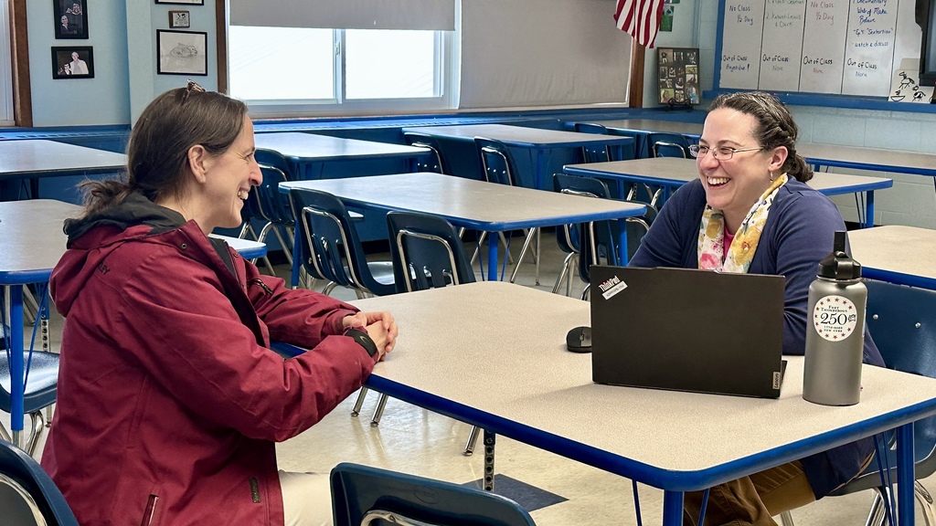 An educator sits across from a parent in a classroom, smiling and talking during a conference with a laptop open on the table.
