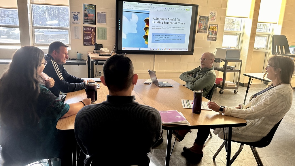 A small group of teachers sit around a table discussing classroom strategies while a presentation about guiding student AI usage appears on a screen behind them.