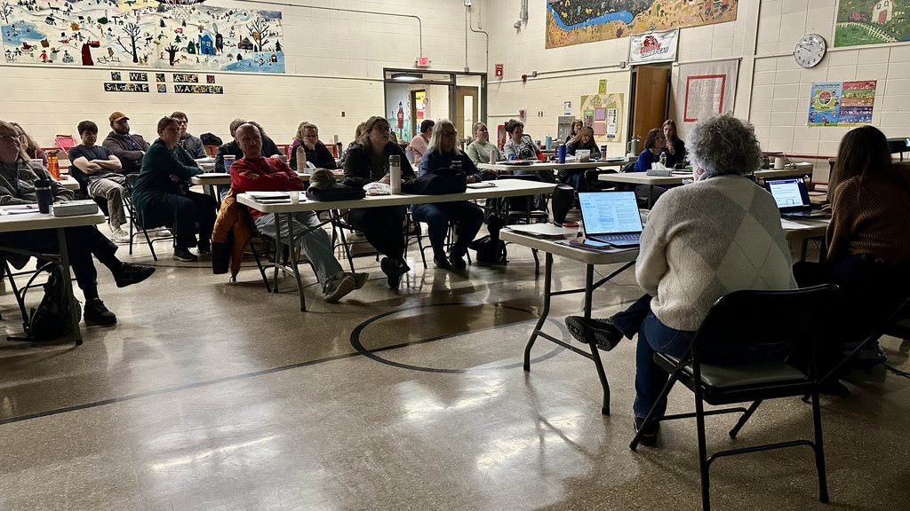 Educators sit at tables in a school gymnasium listening to a presentation during a large-group professional development meeting.