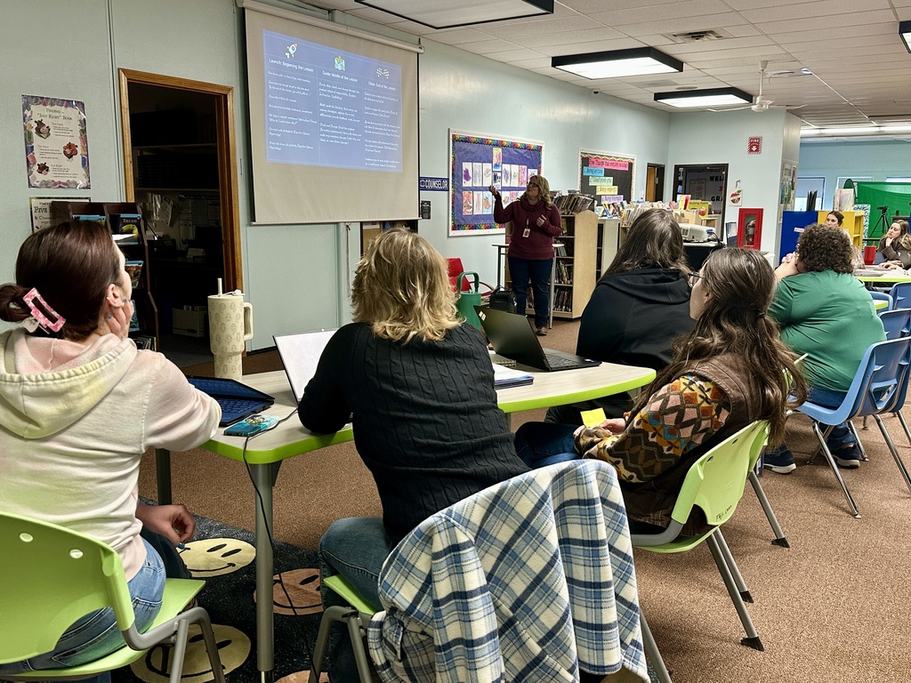 A teacher stands at the front of a classroom pointing to a projected slide while educators seated at tables listen and participate in a professional development workshop.