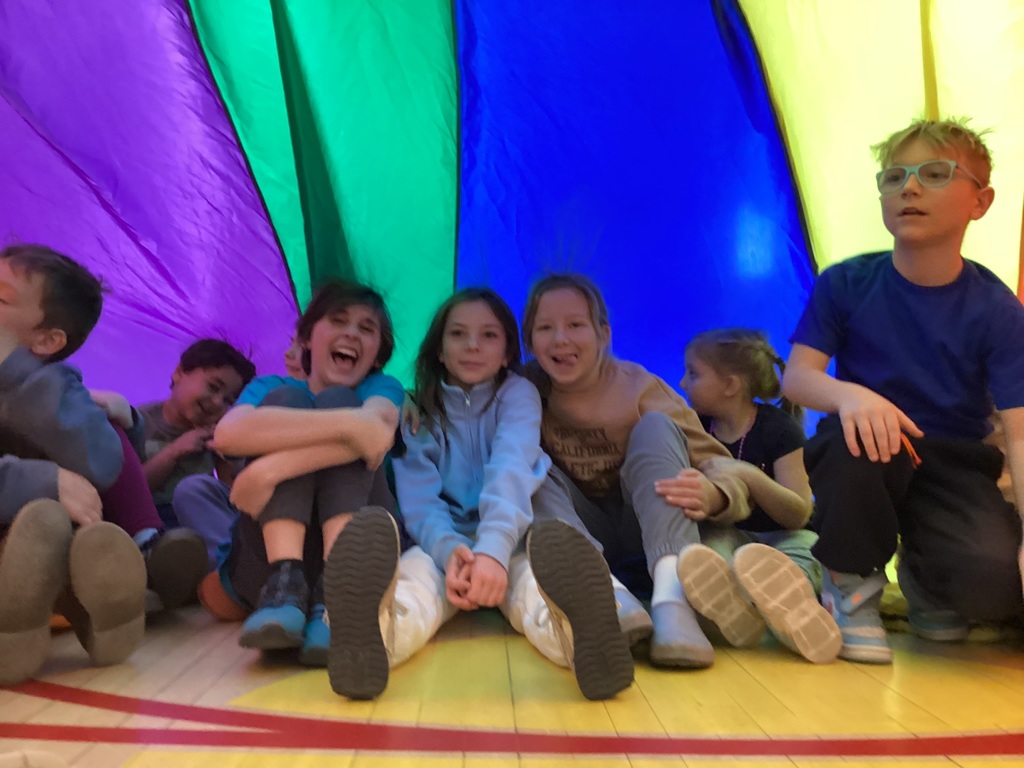 A group of students sit together under a rainbow parachute canopy in a gym, smiling and laughing during a cooperative class activity.