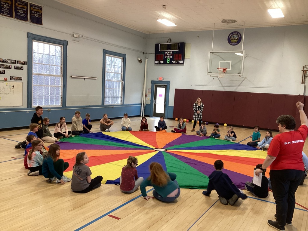 Students sit in a circle on a gym floor around a large colorful parachute while a teacher gives instructions during a group activity.