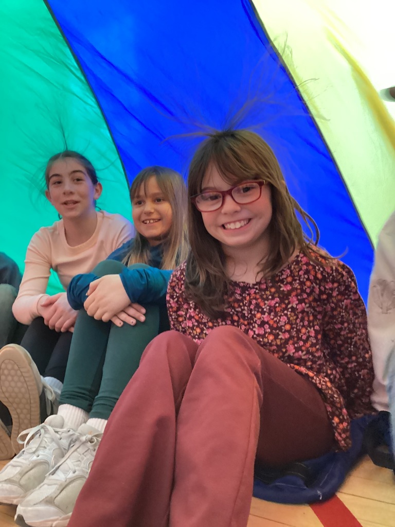 Three students sit inside a raised rainbow parachute in a gym, smiling while the fabric forms a colorful canopy around them.