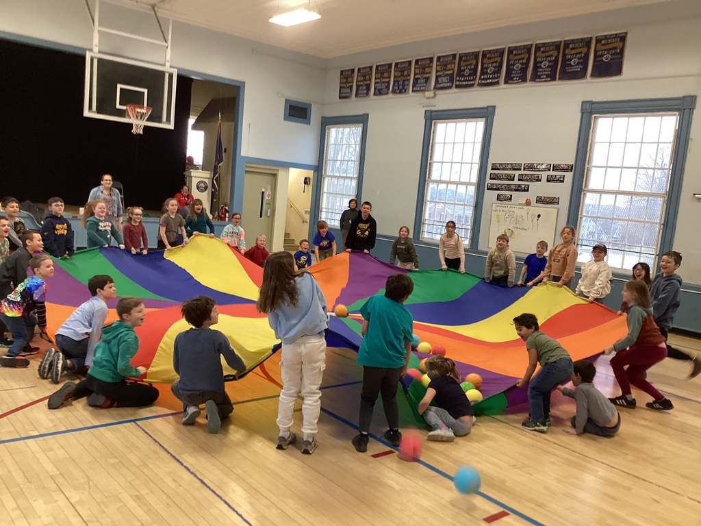 Students in a gym class lift and move a large rainbow parachute together while colorful balls bounce across the surface.