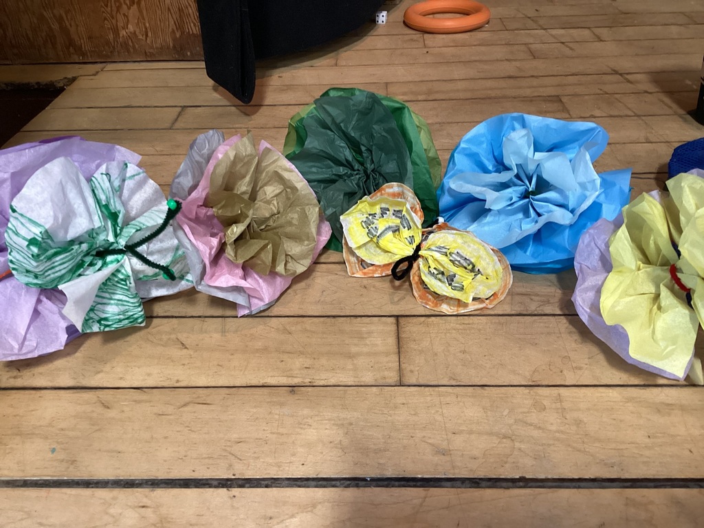 Colorful tissue paper flowers and a handmade paper butterfly displayed on a wooden floor, likely part of a student art or craft activity.