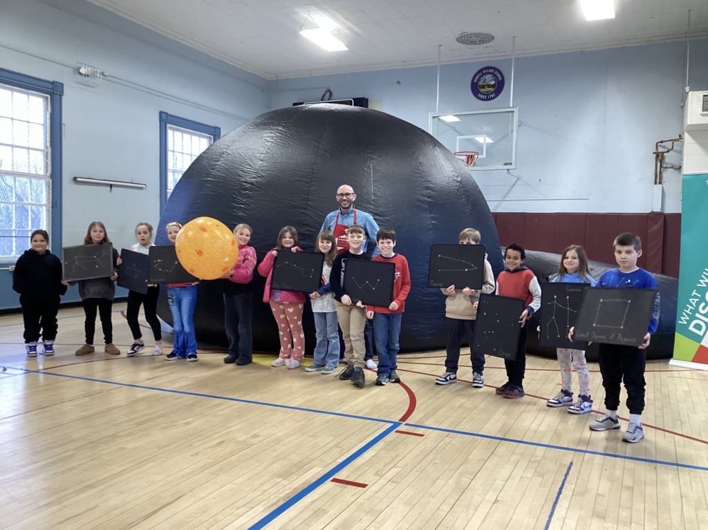 Group of elementary students stand in a gym with a science educator in front of a large inflatable planetarium dome, holding black boards showing constellation patterns.