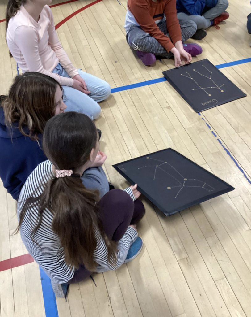 Students seated on the gym floor examine black boards displaying constellation patterns made with connected star points.