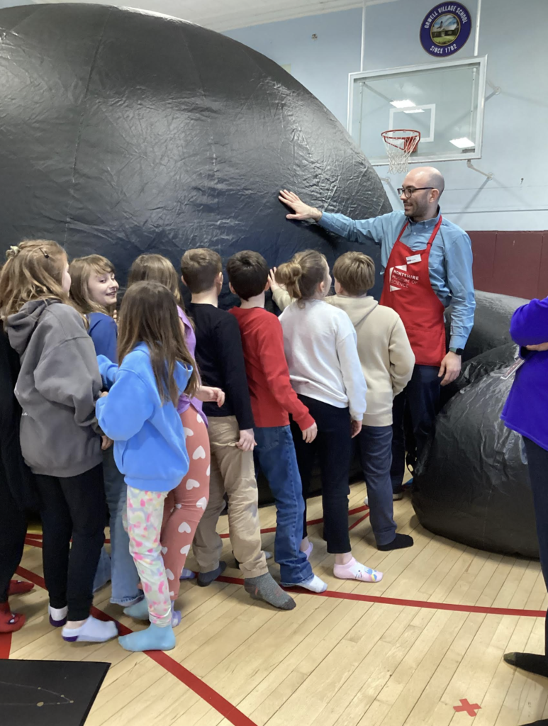Students gather around a science educator in a gym as he points to the large inflatable planetarium dome used for a space and astronomy lesson.