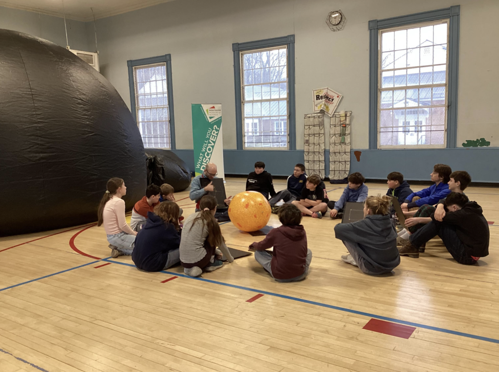 Students sit in a circle on the gym floor around a large orange model of the sun while a science educator leads a discussion during a space science activity.