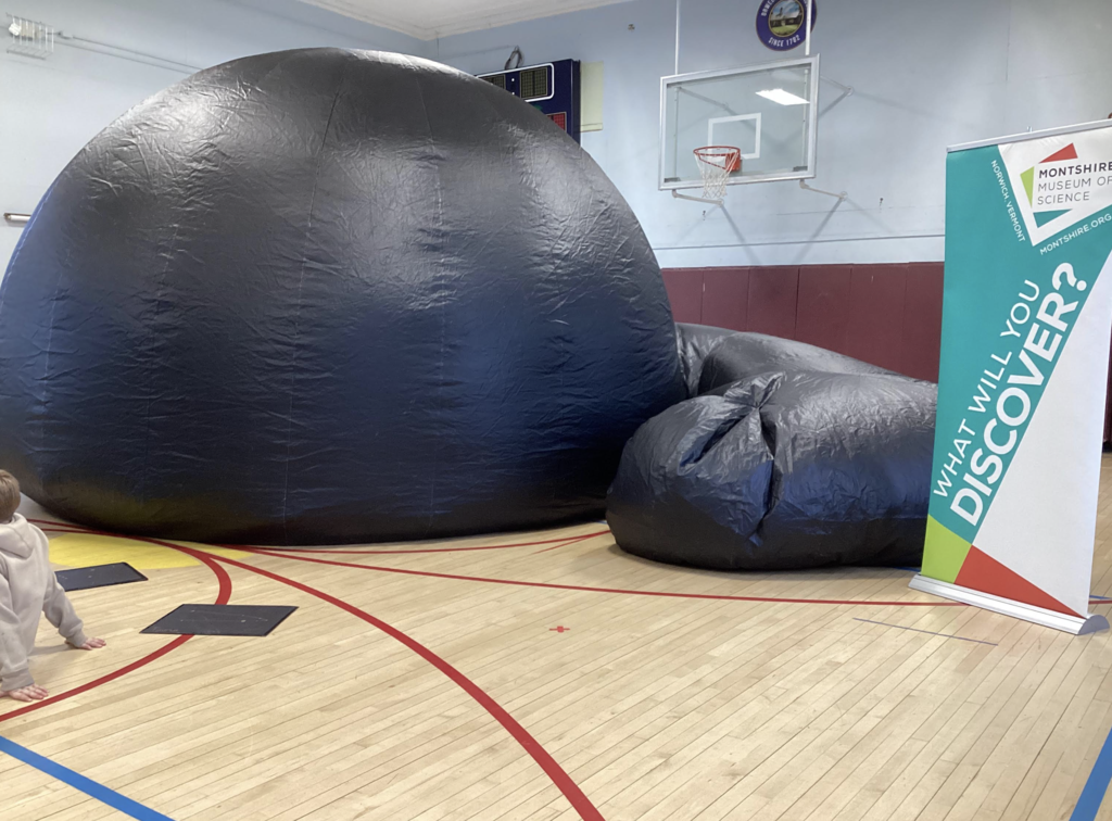 Large inflatable planetarium dome set up in a school gym beside a Montshire Museum of Science banner reading “What Will You Discover?”**