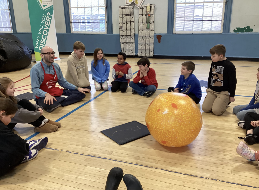 Science educator sits with students in a circle around a large orange sun model, discussing constellations and space concepts.
