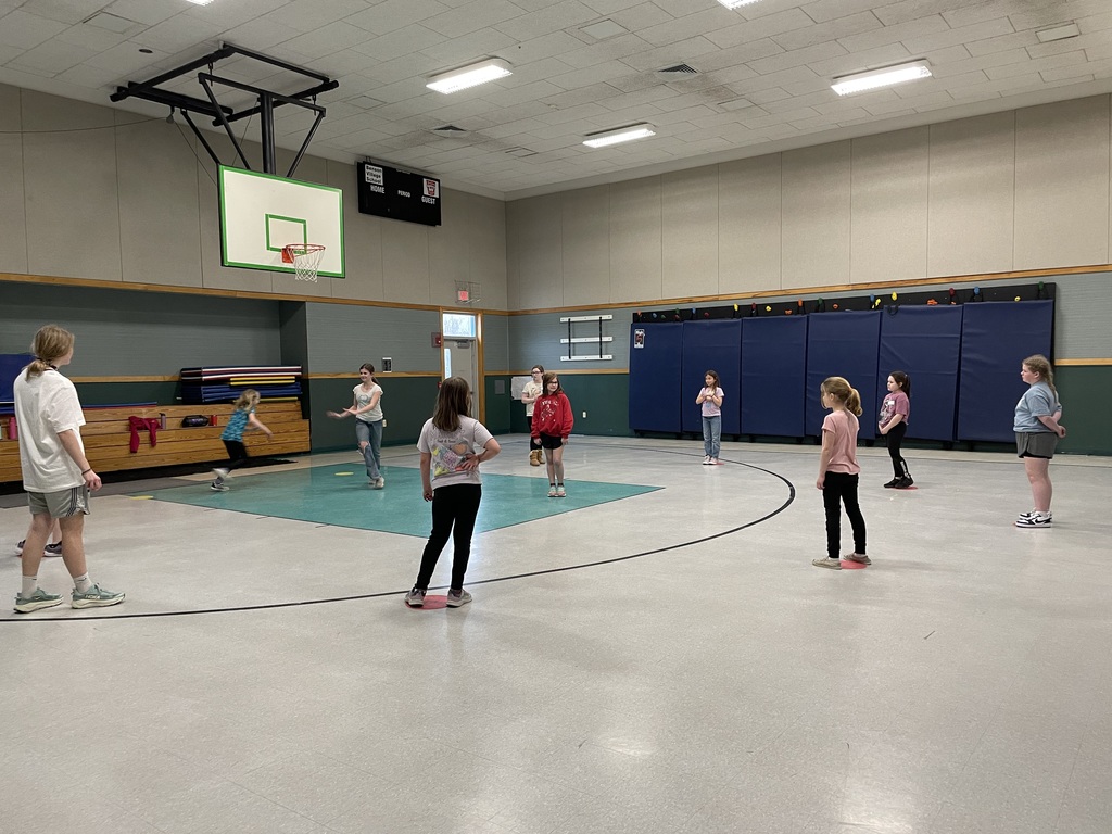 A medium outdoor wide shot features a high ceiling gymnasium as a crowd of eight young girls are on the court. On the left side of the gym are four girls standing on the court. One is wearing a light grey long-sleeved shirt with shorts and grey shoes. The person is facing right towards another girl on her left, who is standing with her hands on her hips wearing a white short-sleeved shirt, black pants, and red shoes while facing backwards. Another girl next to her is wearing a red long-sleeved shirt with black pants. In front of her, facing the person, is a girl standing wearing a beige short-sleeved shirt with blue pants. Behind these four girls, on the far left, is a tall girl who is wearing a white short-sleeved shirt, light beige shorts, and light grey shoes. She is facing right with her hands at her side.  On the right side of the gym are four other girls who are wearing:  Short-sleeved pink shirt, dark blue pants, white shoes facing forward.  Long-sleeved pink shirt, black pants, and pink and white sneakers, facing backwards.  Long-sleeved dark purple shirt, black pants, and black sneakers facing forwards.  Short-sleeved light blue shirt, black pants, and white sneakers facing left.  Behind the two girls on the far left are padded horizontal boards on the wall. Behind the four girls on the far right are padded vertical boards on the wall. Behind all the girls is a door and a window on the wall that lets natural daylight light in the gym. Overhead is lighting and a scoreboard. The left side has a basketball net. The gym floors are light grey.