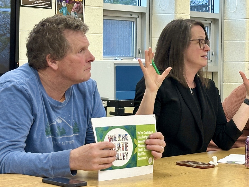 Two adults sit at a table during a meeting, with one person speaking and gesturing while holding a document.
