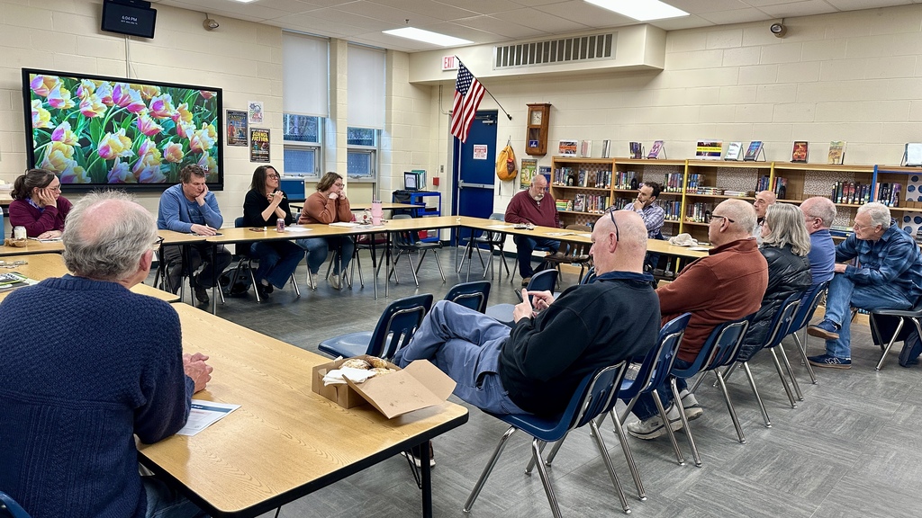 Community members sit around tables in a school library during a meeting, listening and participating in a discussion.