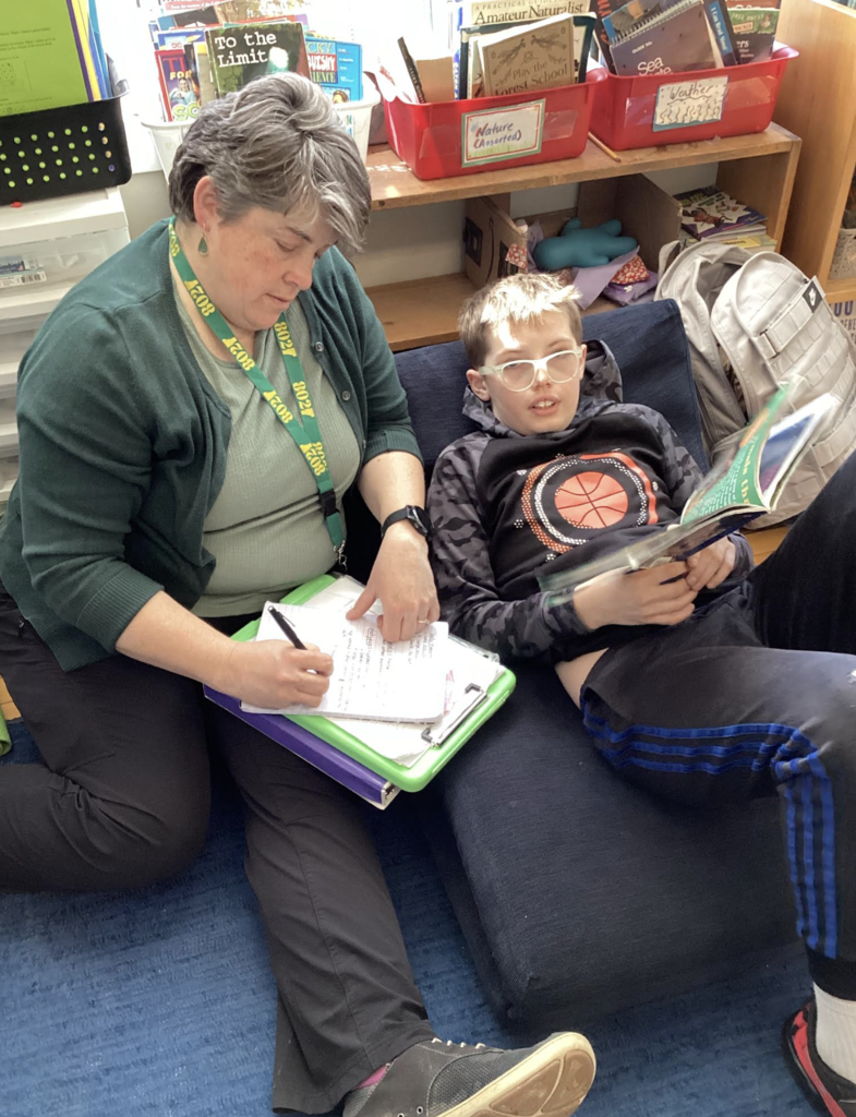 Staff member sits beside a student on a couch, taking notes while the student reads a book.