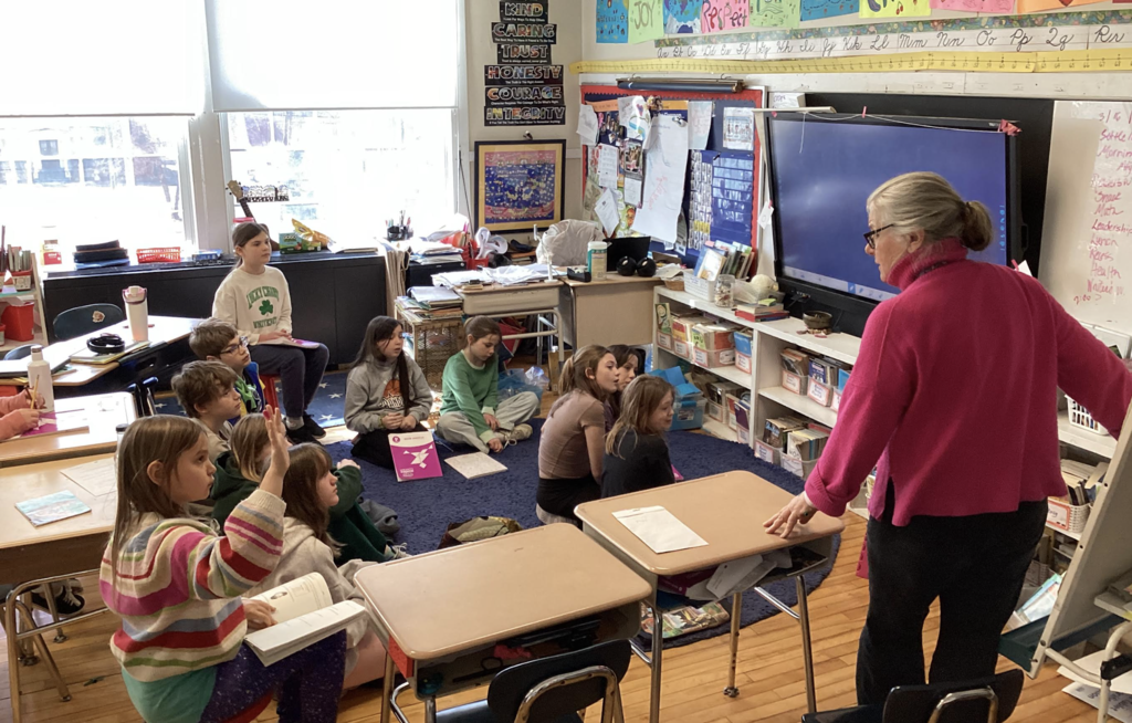 Teacher leads a small group lesson while students sit on the floor and raise hands in a classroom.