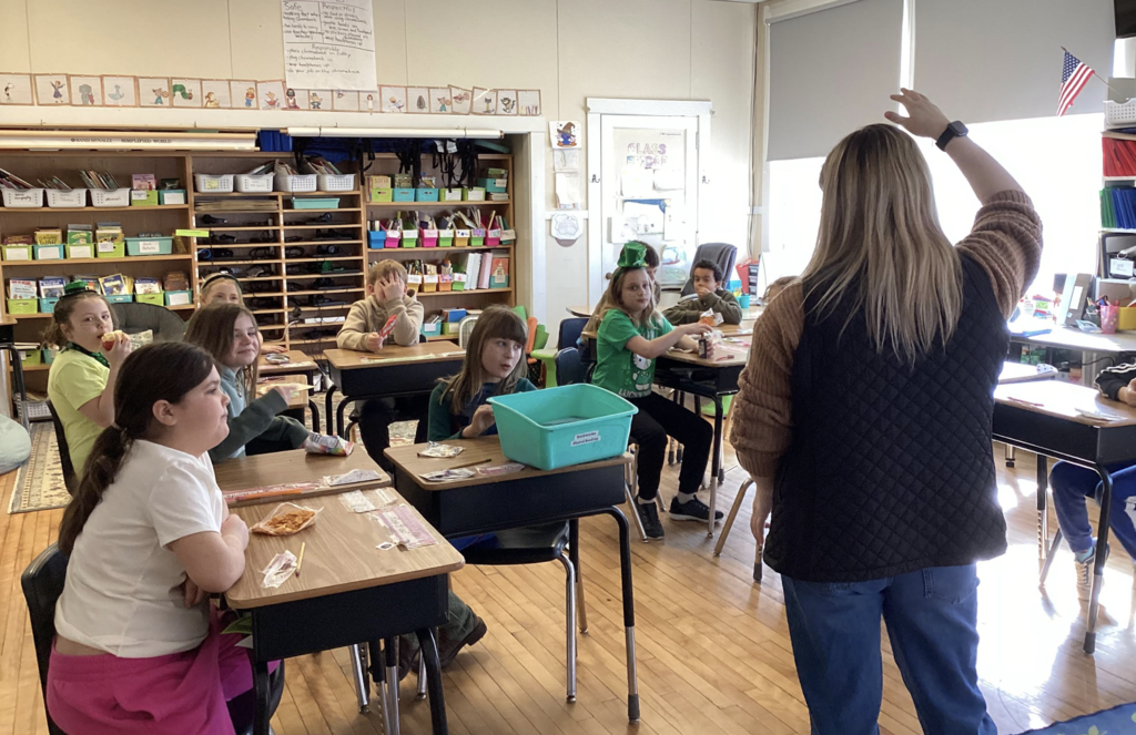 Teacher stands at the front of a classroom with hand raised as students sit at desks and participate.