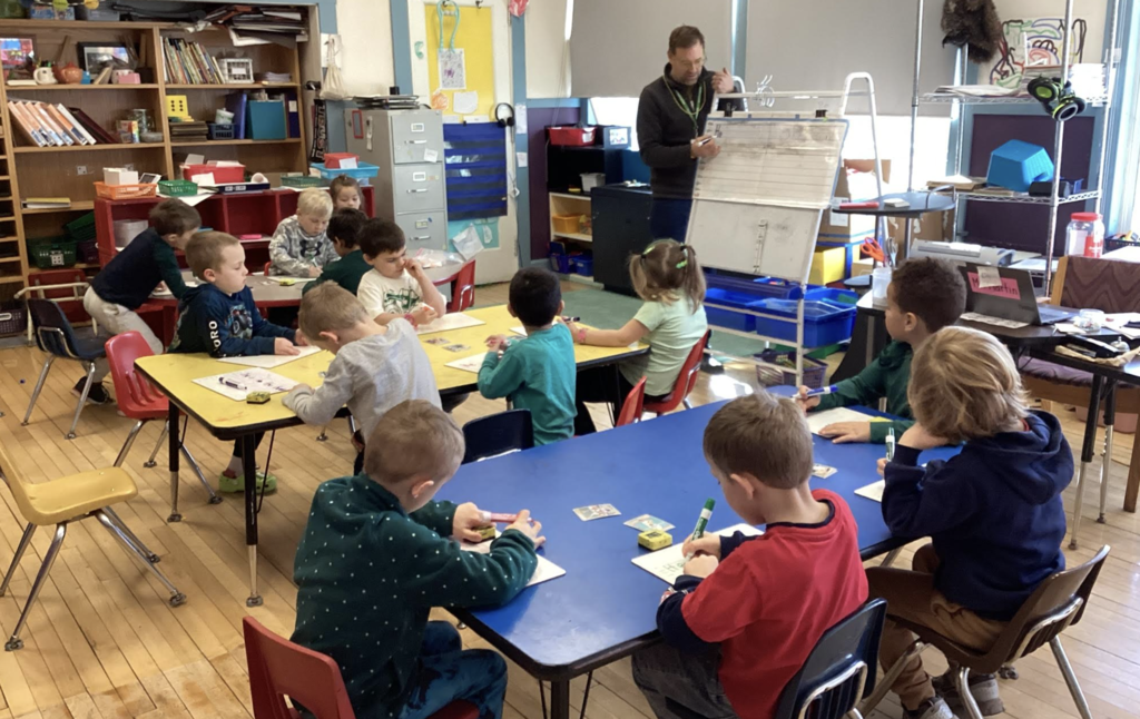 Young students sit at tables writing and drawing while a teacher stands at the front.