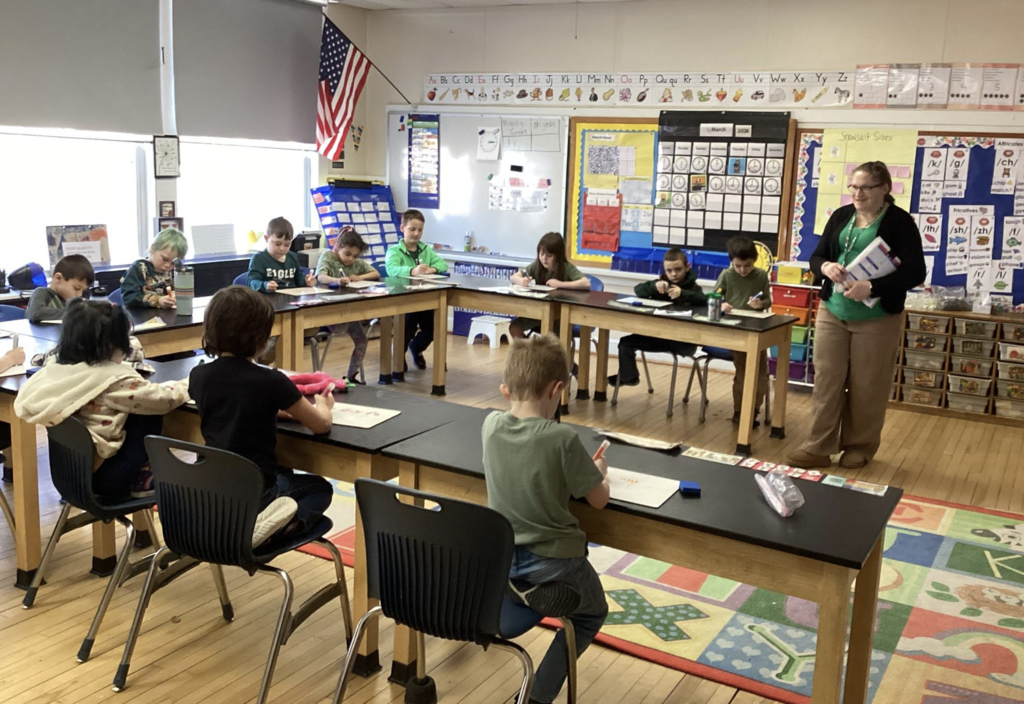 Teacher leads a class as students sit around tables writing on whiteboards.