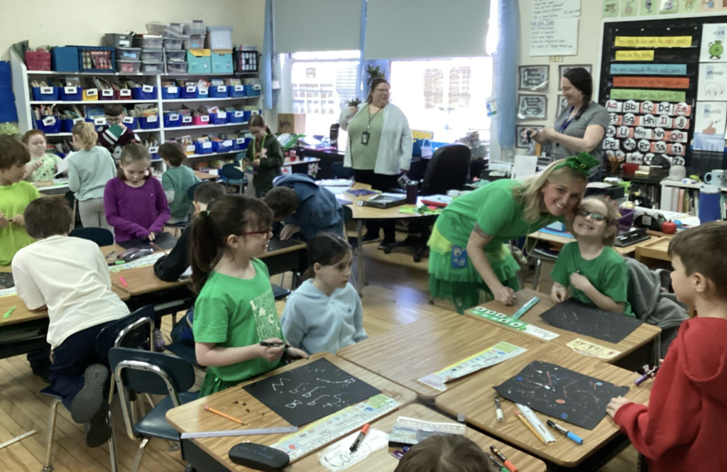 Students and teacher engage in a hands-on classroom activity, creating projects at their desks.