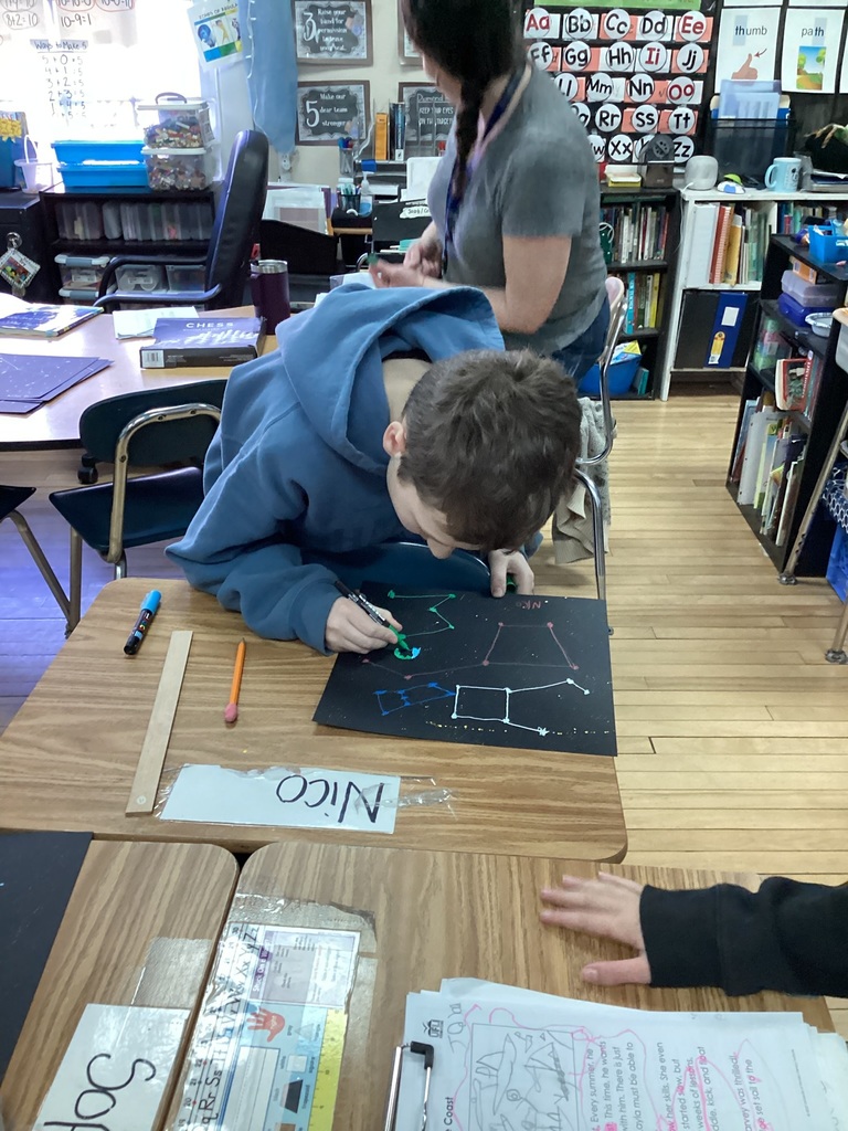 Student works on a constellation drawing at a desk while a teacher sits nearby.