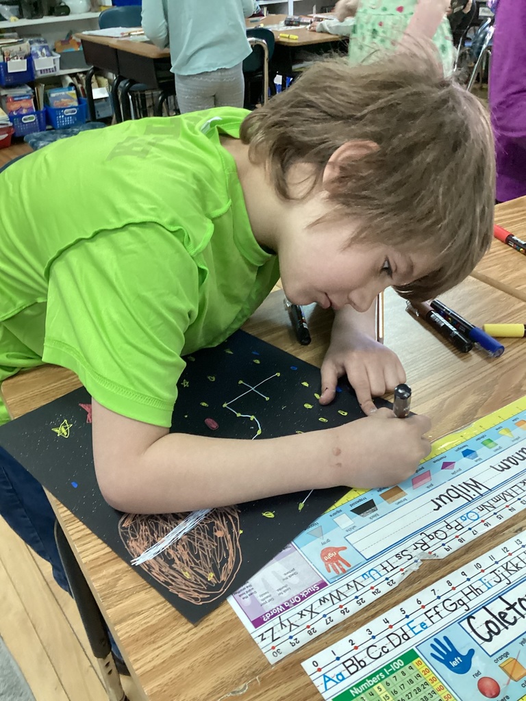 Student leans over a desk, carefully drawing a constellation design on black paper.