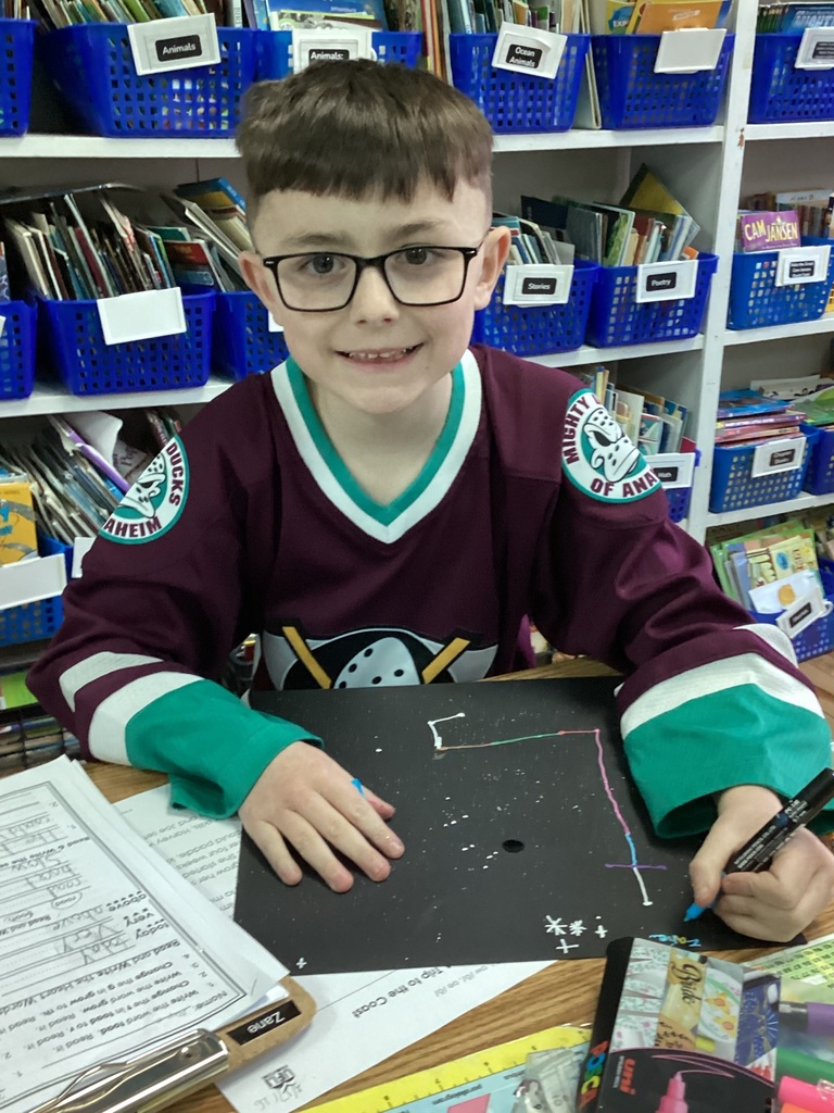 Student wearing glasses and a hockey jersey draws a constellation design at a desk.