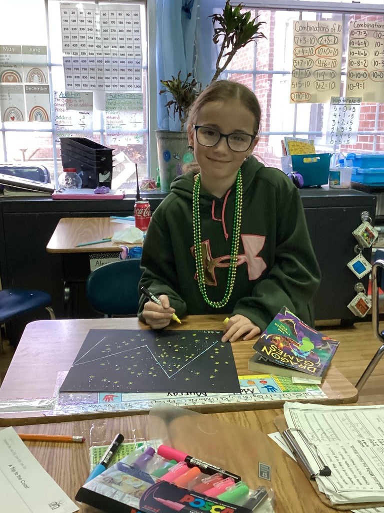 Student wearing green beads smiles at a desk while working on a constellation art project.
