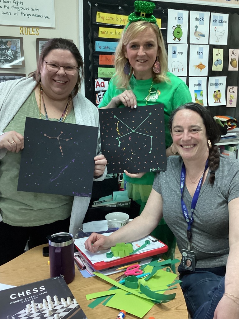 Three staff members smile while holding up constellation-themed student artwork in a classroom.