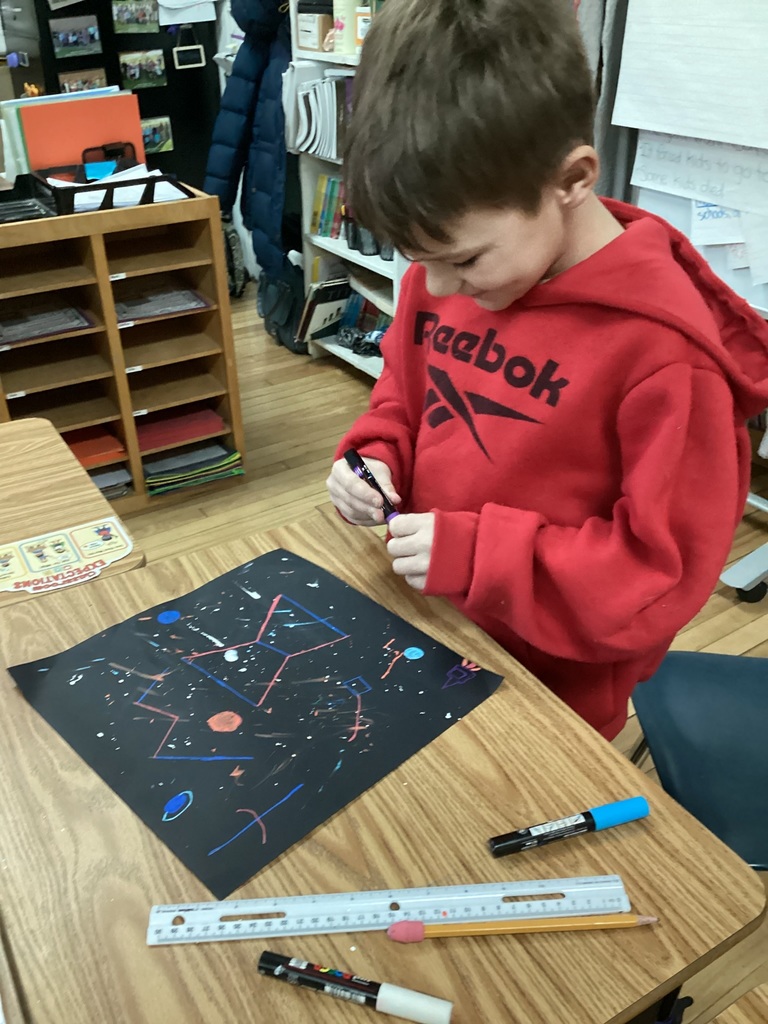 Student in a red sweatshirt draws on black paper with colorful markers at a classroom desk.