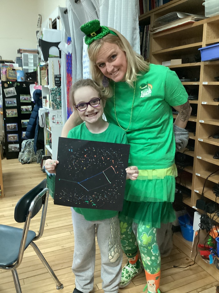 Student and teacher in green attire smile while holding a constellation-themed art project in a classroom.