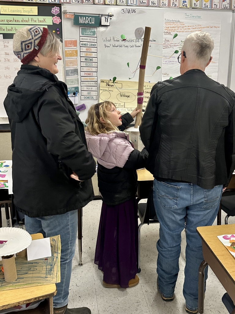 A young student stands between two adults in a classroom, explaining a tall cardboard marble run model while pointing to different parts of the structure.