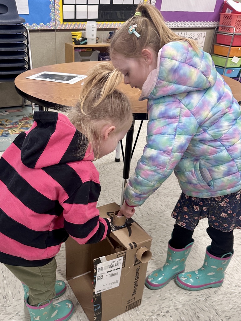 Two students stand over a cardboard structure, dropping a marble into the top of their marble run to test how it travels through the design.