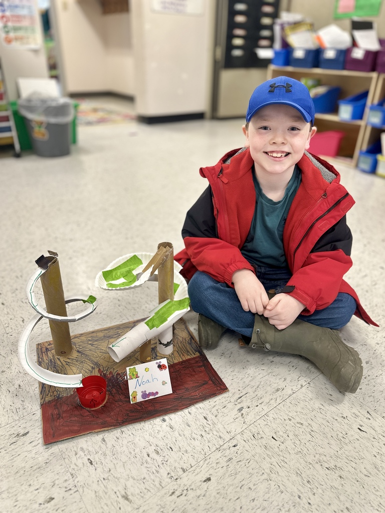 A young student sits on the floor next to a completed marble run made from cardboard tubes and paper plates, smiling proudly.