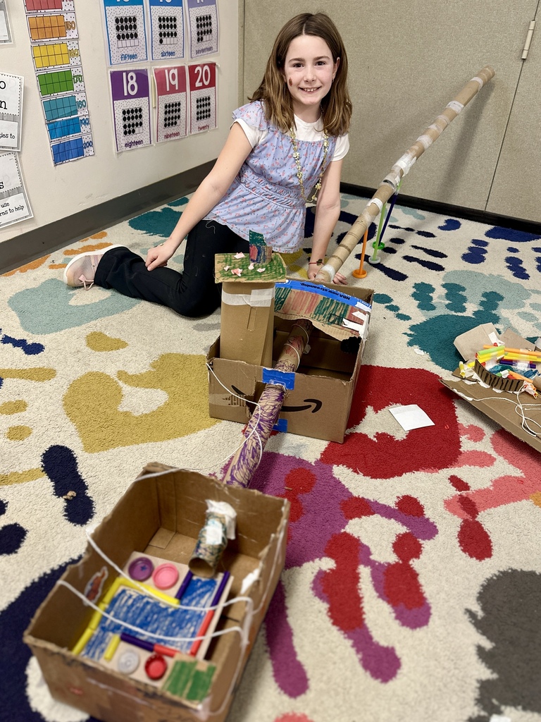 A student kneels on a colorful classroom rug beside a handmade marble run constructed from cardboard boxes and tubes, smiling at the camera.