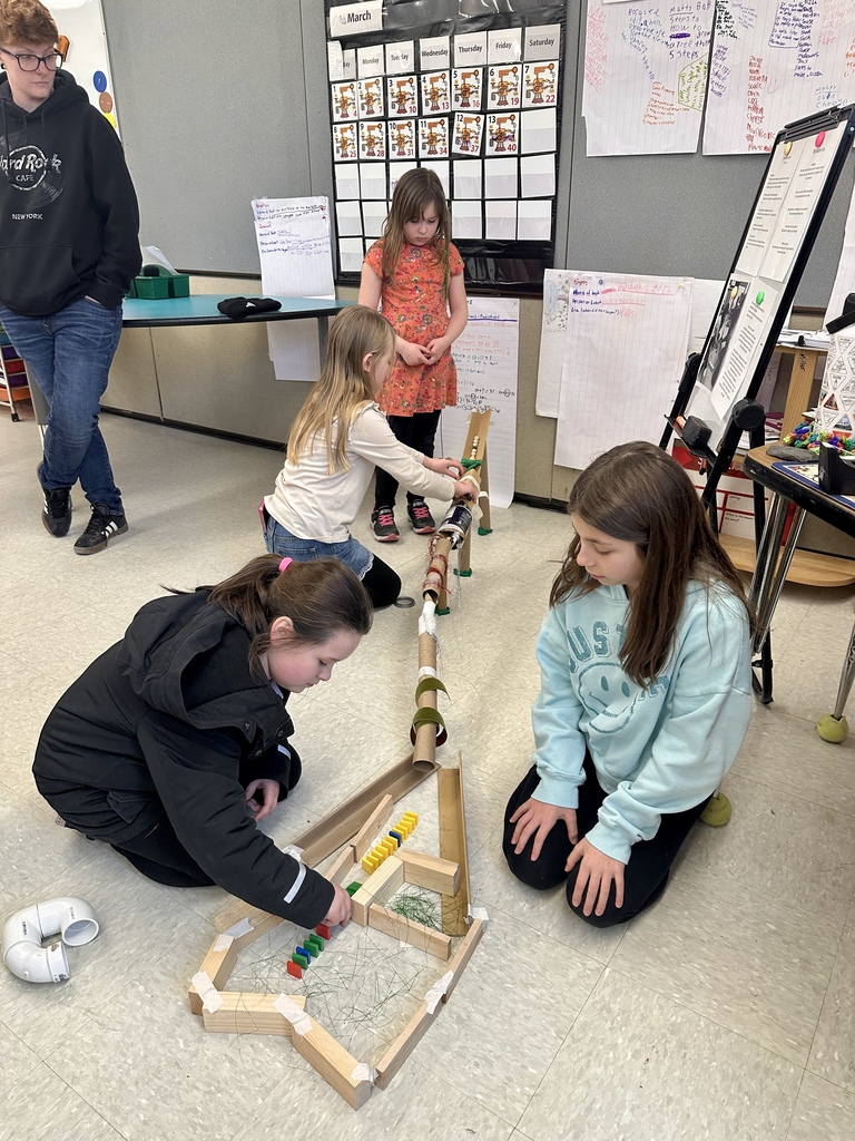 Three students kneel on the floor collaborating on a long marble run made of cardboard tubes, wooden blocks, and other materials.