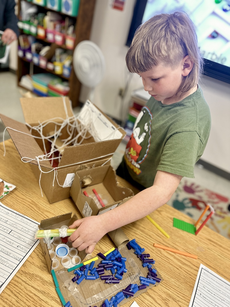 A student stands at a desk working with cardboard, straws, and small plastic pieces while building a marble run during a classroom activity.
