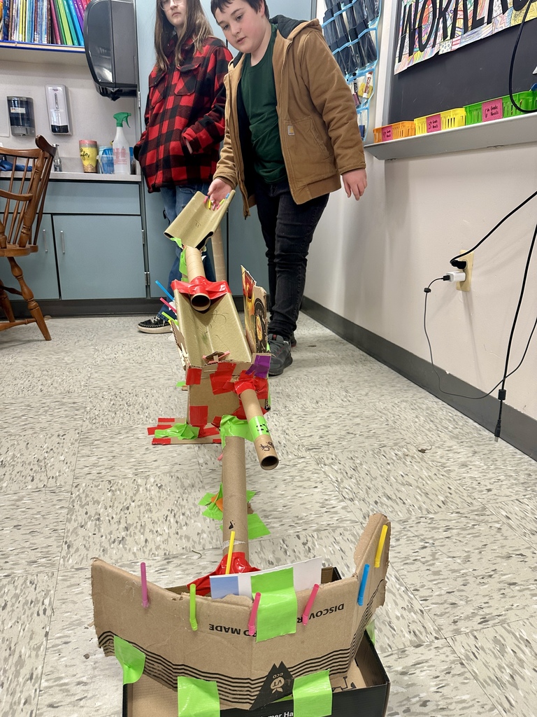 Two students stand beside an extended marble run built along the floor, guiding a marble through the structure during a demonstration.