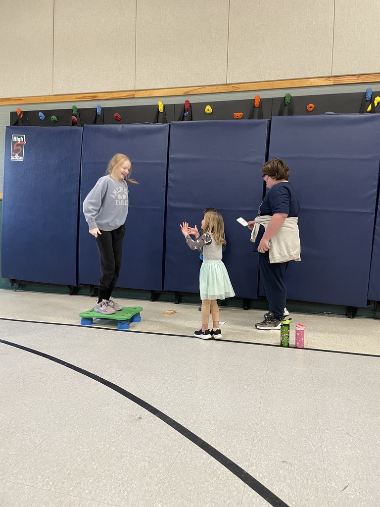 In a gym with blue wall mats, a girl in a grey sweatshirt stands on a small green raised platform. Another girl in a tutu and an older student holding flashcards look on, suggesting a "step-up" or jumping game based on the cards. 