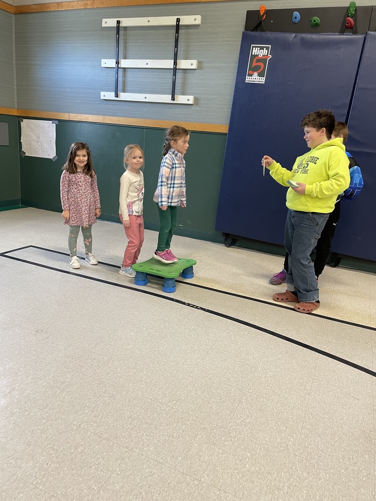  A line of young children waits their turn to jump onto a green platform in a gym. A boy in a bright neon yellow hoodie stands to the side, acting as the leader and holding up a small card for the next student in line.  IMG_6113.jpg: A boy in a red shirt and plaid flannel runs across a gym floor toward a large cream-colored poster on the ground. The poster features various hand-drawn letters, and several 3D plastic letters are scattered across it for a matching game.  IMG_6119.jpg: Two children are captured in motion playing a letter-matching game in a gym. A girl in a tutu skips toward the camera while a boy in a blue shirt runs toward the back wall. Between them lies a large paper on the floor covered in colorful alphabet letters.
