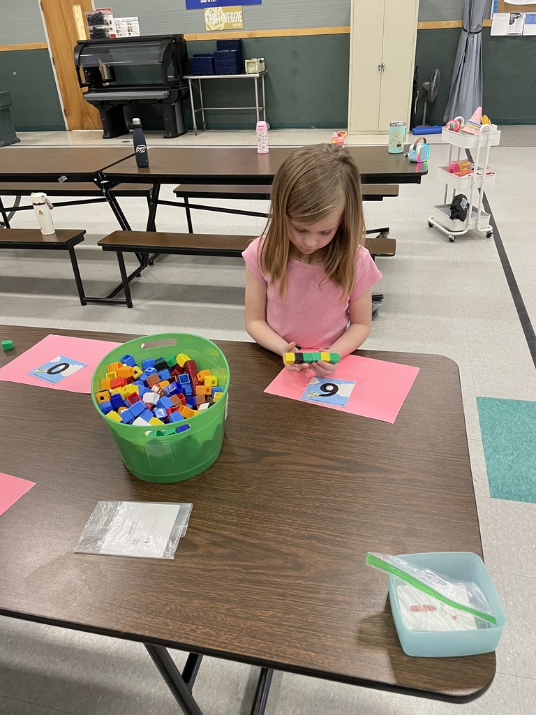 A young girl in a pink shirt focuses on a math task at a table. She is holding a small stack of green and black plastic cubes, sitting in front of a pink paper with the number 6 on it. A large green bucket filled with colorful cubes sits nearby.  Active Learning Games IMG_6114.jpg: In a gym with blue wall mats, a girl in a grey sweatshirt stands on a small green raised platform. Another girl in a tutu and an older student holding flashcards look on, suggesting a "step-up" or jumping game based on the cards.  