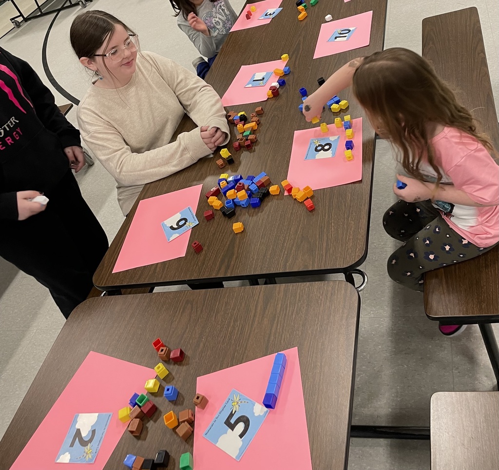  High-angle shot of children sitting at a brown folding table practicing math. They are using colorful interlocking plastic cubes to match the numbers printed on blue sky-themed cards (like 2, 5, 8, and 9) placed on pink sheets of paper. 