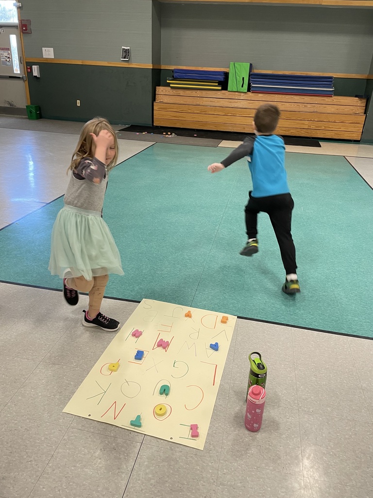 : Two children are captured in motion playing a letter-matching game in a gym. A girl in a tutu skips toward the camera while a boy in a blue shirt runs toward the back wall. Between them lies a large paper on the floor covered in colorful alphabet letters.