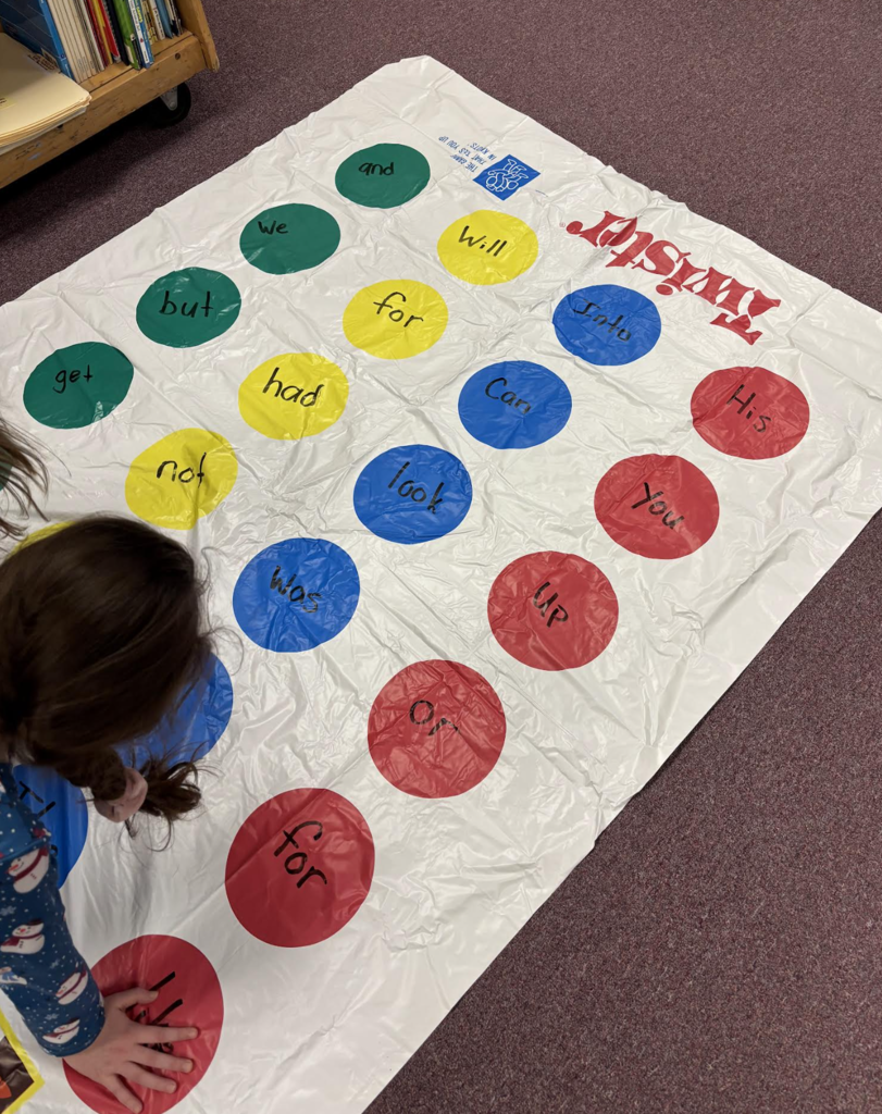 A Twister-style mat on the floor labeled with sight words in different colors, used for a literacy-based game activity.