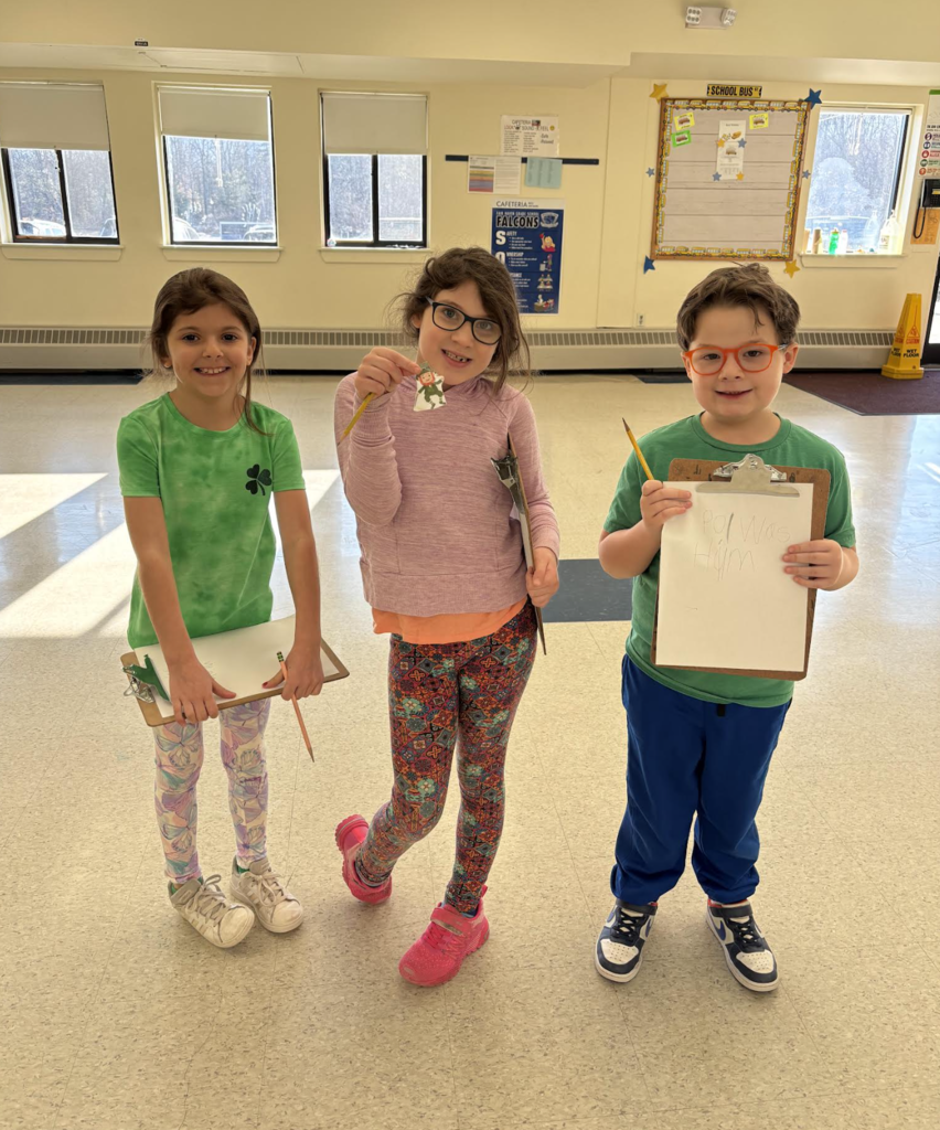 Three elementary students stand in a school hallway holding clipboards and pencils, smiling during a classroom scavenger hunt activity.