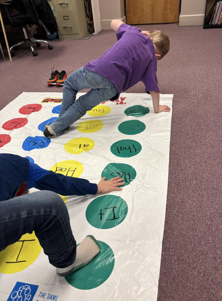 Students play a Twister-style word game, reaching for colored circles labeled with sight words while practicing reading skills.