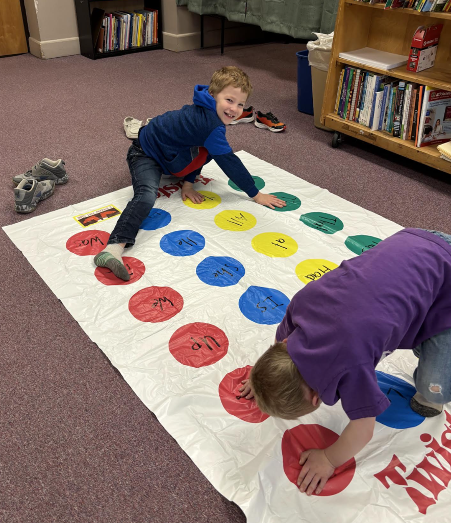 Two students play a Twister-style game with sight words, balancing on colored circles while smiling and engaging in the activity.