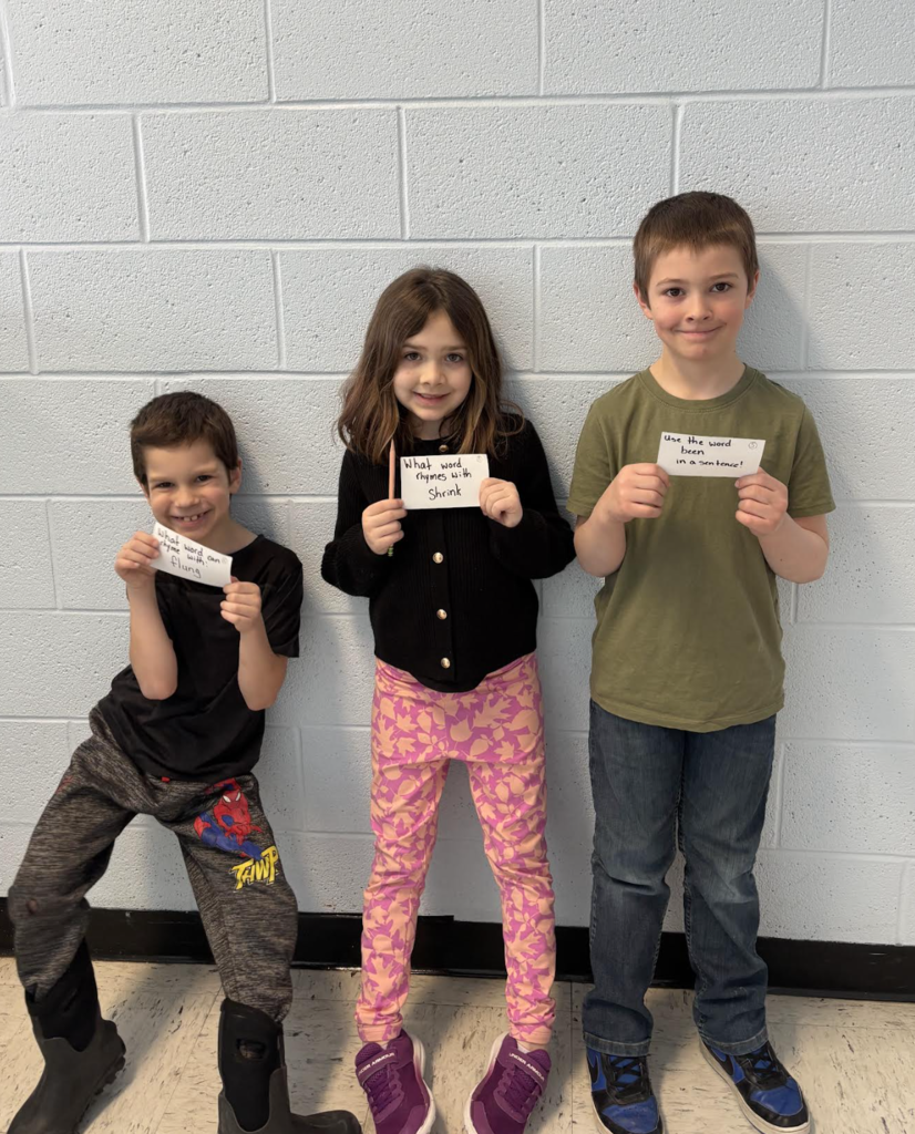 Three students stand against a wall holding cards with reading prompts, participating in a literacy scavenger hunt.
