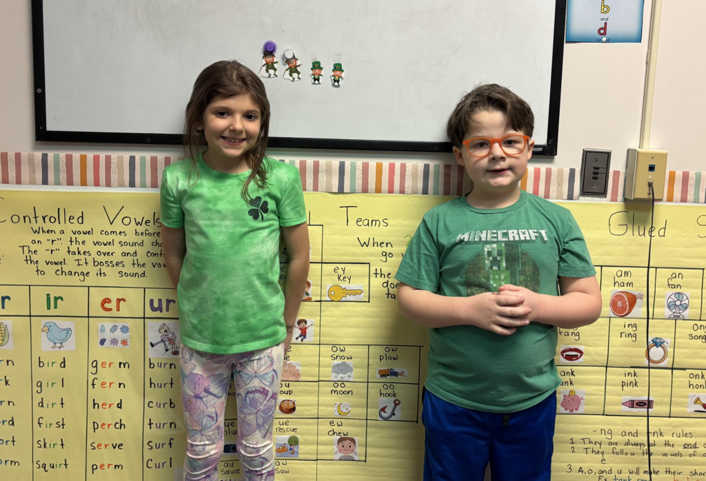 Two students stand in front of a classroom poster about vowel sounds, smiling during a literacy activity.